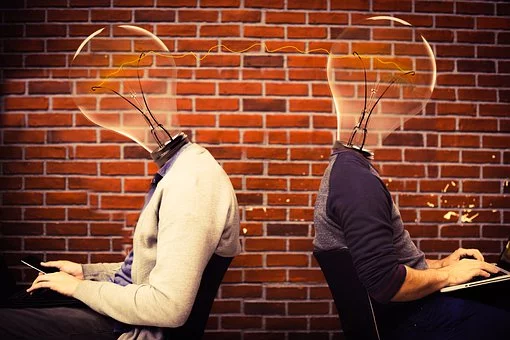 La imagen muestra dos personas trabajando con dos portátiles pero en lugar de sus cabezas tienen bombillas