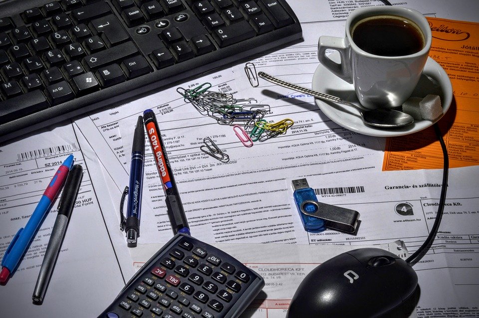 La imagen muestra una mesa de trabajo llena de papeles, bolígrafos, clips, usb y un café